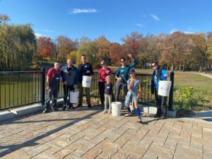 8 individuals stand on a concrete title platform infront of a background of a green pond and multicolroed trees. The sky is bright blue and individuals are dressed for cool weather.